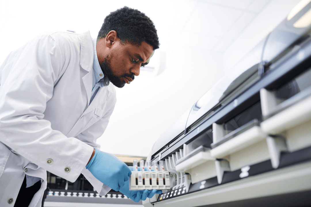 A scientist in a white lab coat and blue gloves loads a rack of sample tubes into a laboratory analyser.