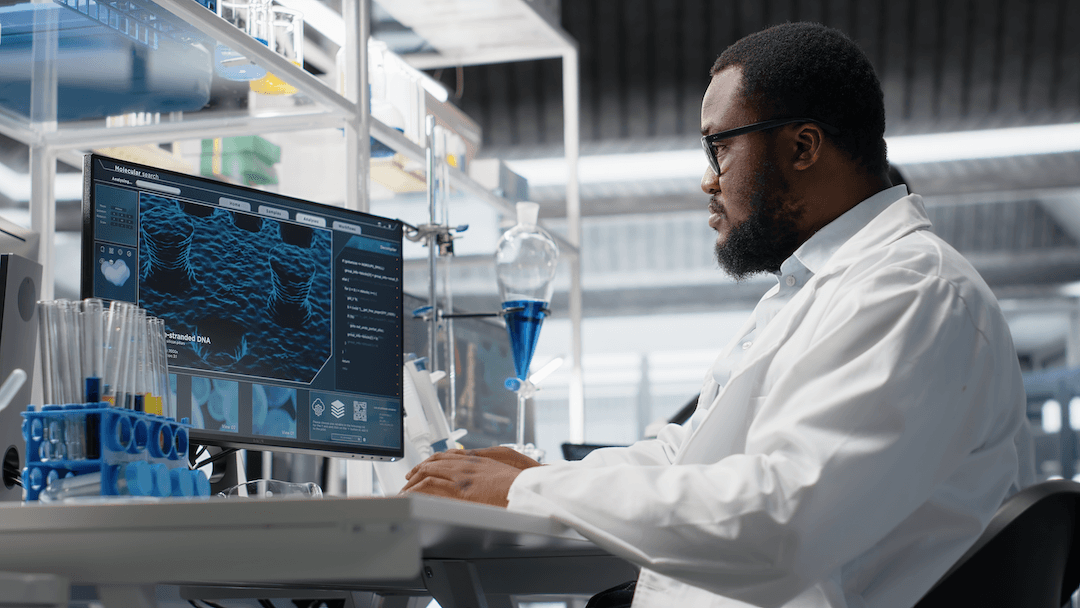 A scientist in a lab coat and glasses works at a computer showing a DNA visualisation in a laboratory.